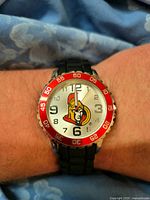 Close-up front view of the watch face showing the Ottawa Senators logo, red bezel with minute markers, black numbered dial, and black segmented strap.