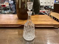 Clear crystal glass decanter with intricate pinwheel cut pattern and stopper, positioned on wooden surface with background showing part of a wooden table and objects.