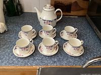 Teapot with six matching cups and six saucers arranged on countertop showing floral design and condition.