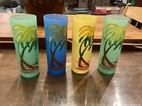 Four tall frosted glasses in green, blue, yellow, and light green colors with a palm tree and sailboat painted design