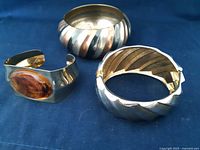 Three bracelets on a blue fabric background showing the tri-color wide band, the gold-tone cuff with faux amber stone, and the gold-tone hinged bracelet.