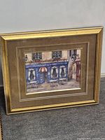 Framed painting showing quaint outdoor café with blue storefront and tables under a framed glass.