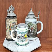 Three porcelain beer steins on white marble pedestal tray. Beer steins have colorful painted scenes with pewter lids.