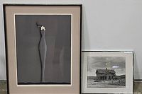 Both framed artworks side by side on the floor against a white wall. One is a tall colored lithograph of a stylized female figure titled "Salomo" by Paul Wunderlich, the other a framed black and white image of a cabin.