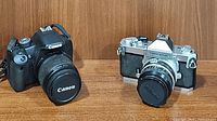 Front view of Canon EOS Rebel T1i 500D DSLR with lens and vintage Nikkormat film camera side by side on a wood surface.