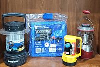 Photo of the entire lot showing the Rayovac lantern, Everbilt tarp in packaging, Eveready flashlight, and GE indoor/outdoor lantern against wooden background.