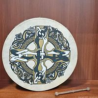 Front view of 18-inch Celtic Bodhran drum with colorful Celtic cross motif on drum skin, resting vertically on a wooden surface next to wooden beater.