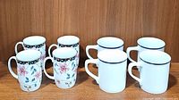 Four Otagiri porcelain floral coffee mugs alongside four white striped coffee mugs, displayed on a wooden surface.