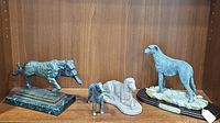 Four dog statues displayed together on a wooden shelf including dog on green marble base, Irish Wolfhound on rocky base, laying dog, and small dog figurine.