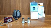 Full view of all items in the lot including opera glasses, Nikon Coolpix L22 red camera, Eagle Optics binoculars, LED night light and Fitbit in packaging.