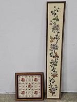 Two framed embroidery art pieces shown upright against a gray wall. One frame is tall and narrow with a floral vine design; the other is a square with a floral grid pattern.