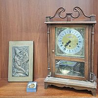 Framed bronze sculpture, vintage brass and wood clock, ammonite pendant box, and brass dog sculpture shown together on shelf.