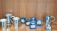 Six metal decanter labels displayed in blue holders, a lidded metal pint tankard, a pair of mint julep cups, a pewter beer mug, and a pewter wine funnel on a wooden shelf.