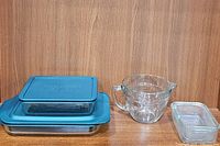 Two covered square Pyrex dishes with blue lids stacked on top of a large rectangular glass dish. Also included is a large clear glass measuring cup and a small rectangular glass dish.