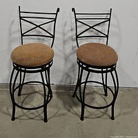 Pair of wrought iron swivel bar stools with brown cushioned seats shown side by side against a white wall and concrete floor.