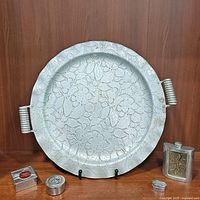 Front view of the round vintage aluminum serving tray with embossed floral pattern and twin handles, accompanied by the metal flask and three metal trinket boxes arranged in front of it.