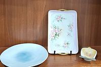 Photo showing the round blue-toned platter on the left, the rectangular floral tray with gold handles upright on a stand, and the small birdcage sponge cradle on the right