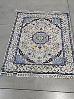 Full view of the rectangular rug lying flat on gray flooring showcasing its floral and geometric blue, green, cream, and black pattern with white fringes at edges.