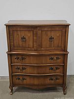 Front view of the wooden tall chest showing 3 drawers and a top cabinet with double doors, featuring ornate metal handles and ring pulls.