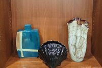 Three vases displayed together: blue ceramic with beige cross, black ceramic shell, and hand blown earth-toned glass handkerchief-shaped vase.