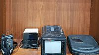 Four vintage electronics items displayed on wooden shelf: small portable Crown black vintage TV, Sony Dream Machine clock radio, black Ambico VHS cassette rewinder, and AIWA cassette player.