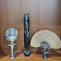 Three African wooden statues displayed on a wooden shelf: one tall slender figure, one small brown fertility statue, and one Ashanti doll with a woven rattan headdress.