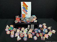 Wide view of over 50 enamel pins arranged on black risers and white board backdrop, showing various shapes and designs.