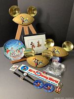 Display of three 35th Anniversary gold Mickey Mouse ears hats with painted Mickey graphics, framed animation cell print with Mickey and Pluto, commemorative plate, two watches, pass lanyards, keychain, and Disney pins arranged on a wooden stand against black background.