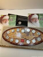 Photo shows Canadian Millennium 1999 coin set arranged in a wooden tray shaped like an oval with a map of Canada background. Coins of different denominations and metal compositions are set in the tray with a clear protective cover. Includes an informational booklet with Santa Claus on cover dated 2008.