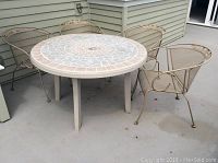 Round plastic outdoor table with mosaic tile look top and surrounding four painted wrought iron chairs with floral leaf design showing outdoor wear.