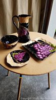 Four-piece Bella Casa serving set displayed on round table including pitcher, square plate, fruit bowl, and stand with black background and grape design.