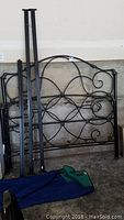 Black metal queen size bed frame headboard, footboard, and side rails stacked in a garage, showing decorative rose and scroll design with some wear and minor scuffs on finish.