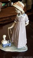 Front angle view of girl in pink dress and hat pulling doll carriage with seated doll inside