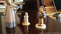 Three porcelain figurines displayed on a table: Nao girl with violin, Goebel Home from Market girl, and Royal Doulton Bedtime Bunnykins bunny.