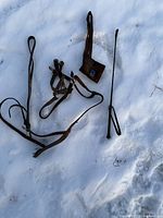 Full view of brown leather horse bridle, black riding crop, and brown suede saddle bag laid out on snow