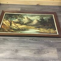 Photo of a framed painting depicting a mountain and stream landscape with trees and rocky terrain on a wooden floor.