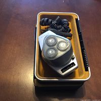 Photo of a silver vintage Philishave HP 1118 electric shaver with three rotary heads, coiled black power cord, and protective plastic cap on a brown tray.