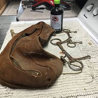 Photo showing a brown suede saddle bag, two metal horse bits, and a bottle of Kopper Kare antiseptic on a textured surface.