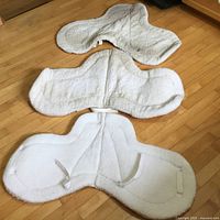 Three white fleece saddle pads of varying sizes laid flat on wood floor showing shape, fabric texture, and securing loops