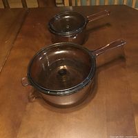 Photo of two brown glass Vision cookware pots with handles and matching lids on a wooden table.