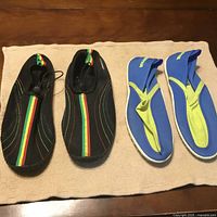 Photo of two pairs of water shoes on a beige textured cloth on wood surface. One pair is black with Jamaican flag colors stripes and elastic closure. The other pair is blue and yellow with a hook-and-loop heel strap.