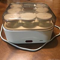 Front view of the Braun yogurt maker showing seven glass jars with white lids under transparent plastic cover. The base is white with Braun branding and a red power light on front.