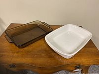 Two rectangular baking dishes on wooden furniture; brown glass Anchor Ovenware and white Corningware