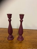 Front view of two burgundy painted wooden candle stick holders with ornate rounded bases and tapering bodies.