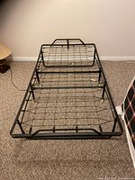 Folding twin metal cot bedframe unfolded on carpeted floor, showing metal wire mesh support surface and foldable joints.
