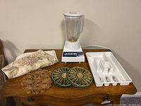 Overview photo displaying all items in the lot on wooden table: Hamilton Beach blender, utensil organizer, trivets, and fabric placemats.