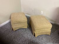 Pair of beige ottoman foot rests placed side-by-side against a wall on a carpeted floor, viewed at an angle.