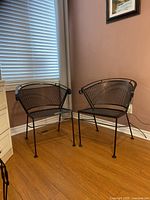 Two black metal patio chairs with curved backs and mesh seats placed on a wooden floor against a beige wall with window blinds.