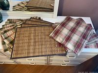 Mixed arrangement of six cloth plaid placemats in red, green, beige and four brown wicker placemats on white dresser.