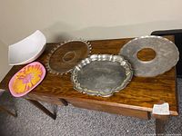 Five serving items displayed on wooden table: large white bowl with chip, pink flower plastic plate, round glass platter, round aluminum tray with center hole, silver-plated raised platter